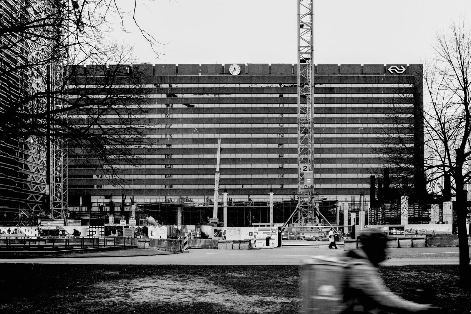 Zwart-witfoto van een gebouw met NS-logo en klok, bouwplaats op de voorgrond, hijskraan en een fietsende bezorger in beeld.