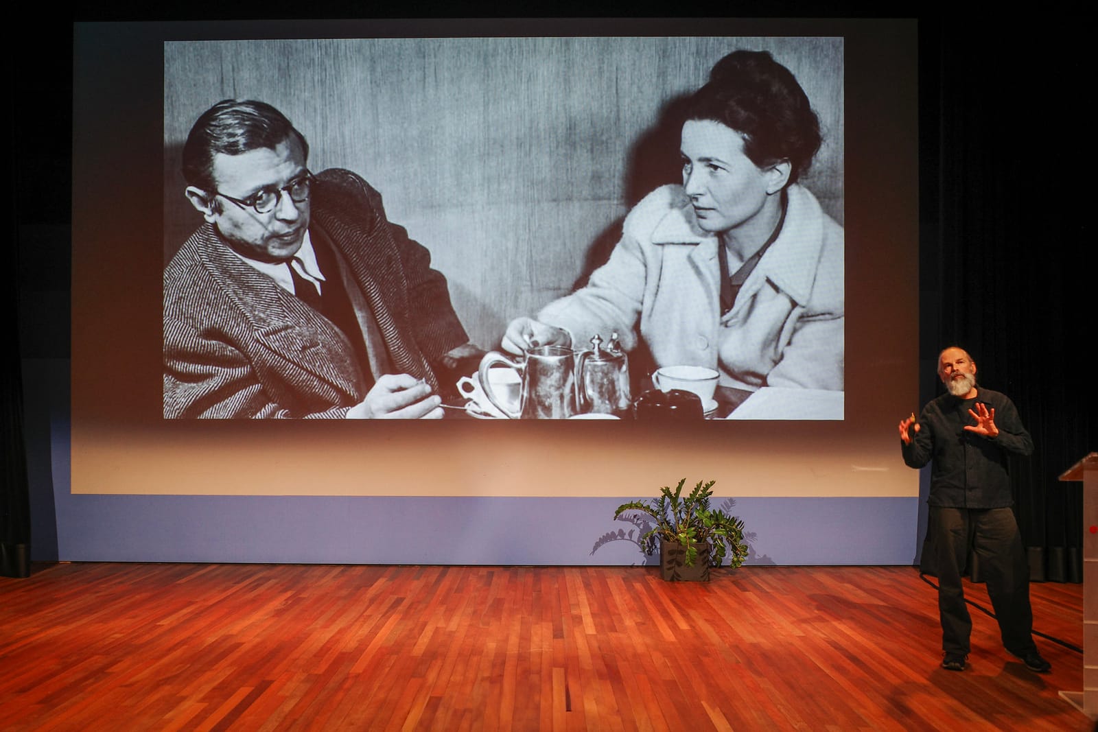 Een spreker presenteert op een podium met een foto van Sartre en De Beauvoir, drinkend, geprojecteerd op een groot scherm.