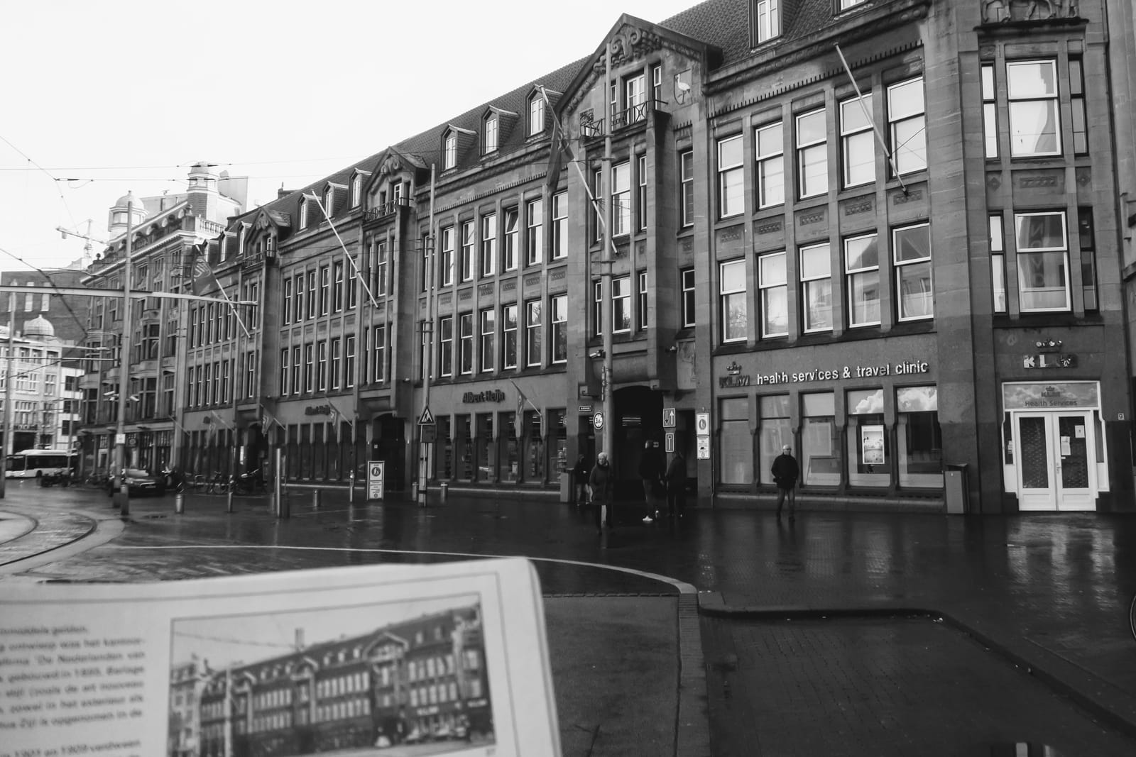 Zwart-witfoto van historische gevels, met winkels en natte straat. Voorgrond toont vergelijkend beeld uit boek.