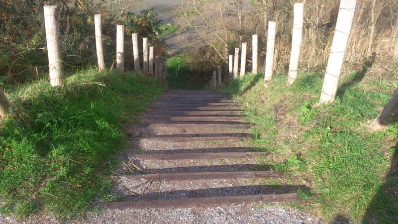 Trap met houten treden tussen groene bermen, omgeven door houten palen, leidt naar beneden in een heuvelachtig landschap.